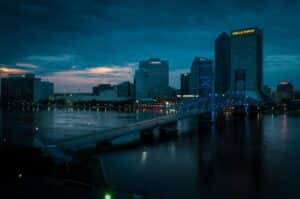 A city skyline at dusk features tall office buildings with illuminated signs and a blue-lit bridge spanning a calm river under a partly cloudy sky with hints of sunset.