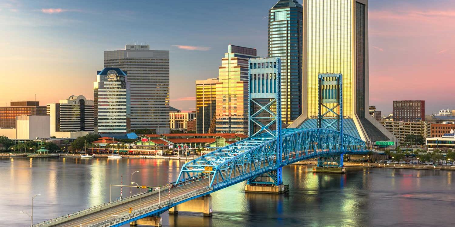 Downtown Jacksonville, Florida skyline at sunset, features modern skyscrapers and the blue Main Street Bridge spanning the St. Johns River, with reflections on the water.