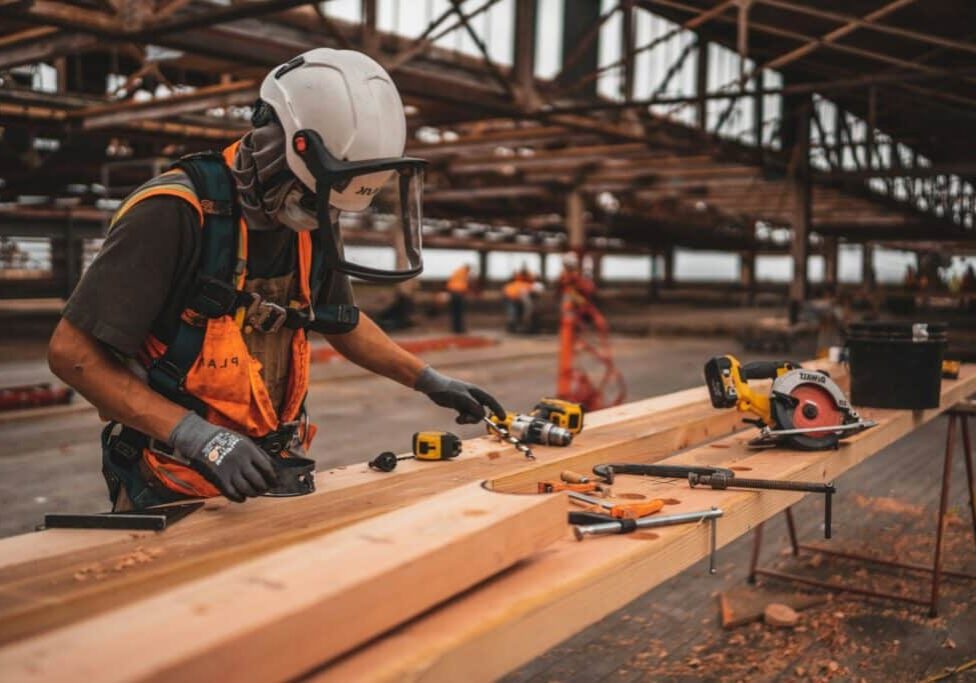 A construction worker wearing safety gear and an orange vest uses tools to work on wooden beams at a job site, with power tools and materials spread out on a workbench in a partially built structure.