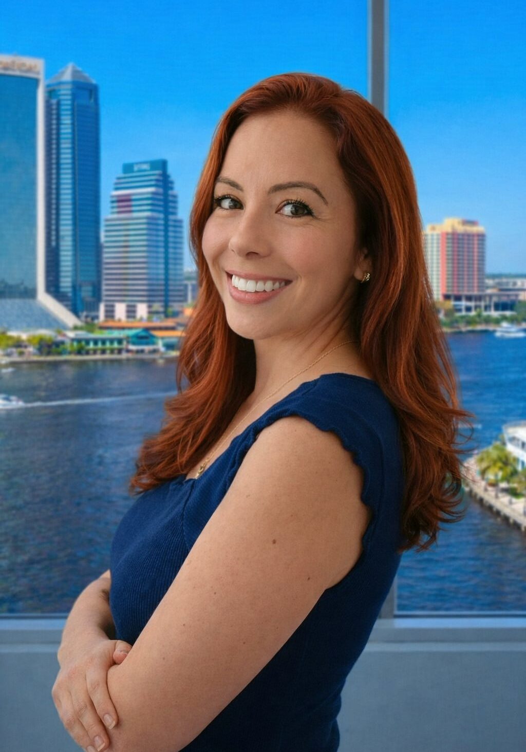 A woman with long red hair, wearing a navy blue sleeveless top, stands smiling with arms crossed in front of a window overlooking a river and a city skyline with tall buildings on a bright, sunny day.